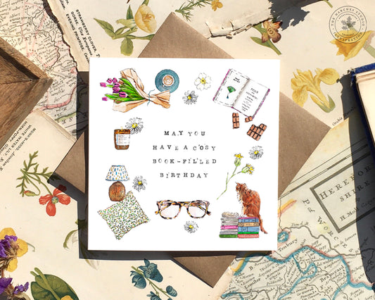 Image of a white square card with delicate illustrations of books, glasses, a cat, and other book lover items. It has 'May You Have a Cosy Book-filled Birthday' in the centre.